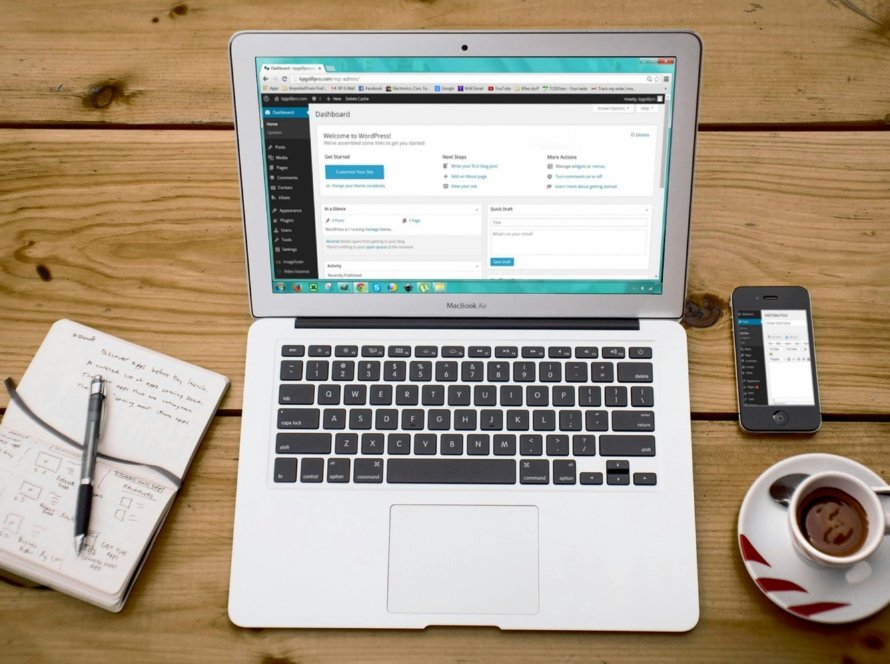 A laptop showing the backend of a website to show WordPress maintenance price in 2024. A coffee cup is on the right and a notepad on the left of the laptop.